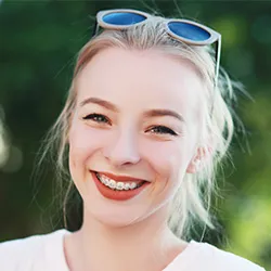 A young woman with braces, blonde hair, and sun glasses smiling outdoors.